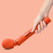 A hand holding the Fun Factory Vim Silicone Weighted Rumbly Wand Vibrator in orange, known for its powerful, deep-reaching vibrations and silicone handle, featuring multiple control buttons. The background is a solid peach color.