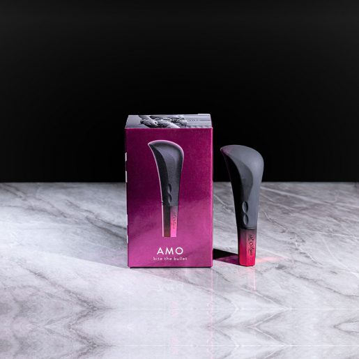 A sleek, black Hot Octopuss Amo Powerful Silicone Bullet massager displayed alongside its matching pink and gray box on a reflective marble surface. The box features an image of the product and the text "Hot Octopuss Amo Bite the Bullet." The photo has a minimalistic, high-contrast aesthetic.