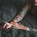 A person with colorful tattoos and red nails is lying on a dark bed, reaching out with one hand holding a Hot Octopuss Digit Silicone Rechargeable Finger Vibrator. The intimate scene is softly lit.