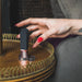 A hand with red-painted nails is holding the Hot Octopuss Digit Silicone Rechargeable Finger Vibrator, an ergonomic toy with a black and gold bullet shape, over a round wooden table with ornate edges. The table features a dark metal ring and a decorative crown emblem nearby. A gray cushion or blanket is partially visible in the background.