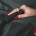 A hand with glossy red nails is holding the Hot Octopuss Digit Silicone Rechargeable Finger Vibrator, a sleek black device equipped with an adjustable finger loop and a small bronze-colored ring. The product is positioned against a dark, textured background.