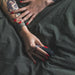 A close-up of two tattooed hands with red nails holding the Hot Octopuss Digit Silicone Rechargeable Finger Vibrator against a dark green fabric background. One hand rests on top of the other, showcasing the tattoos and a ring on a finger.