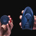 Two hands are shown against a black background, one holding a dark-colored puck and the other holding a Hot Octopuss Pulse Duo Remote Controlled Vibrating Penis Masturbator with remote controlled vibrations, likely a gadget or tool.