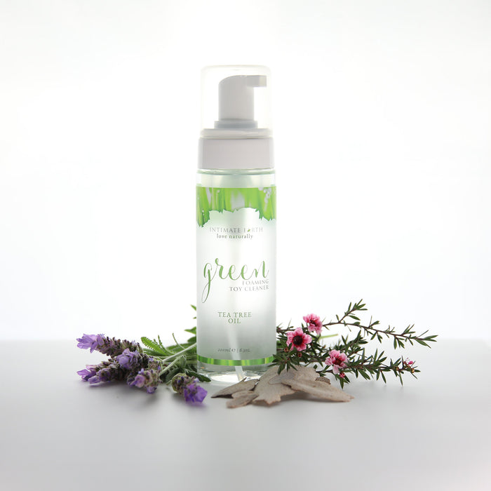 A bottle of Intimate Earth Green Tea Tree Oil Foaming Toy Cleaner 3.4oz stands upright on a white surface, surrounded by sprigs of lavender, pink flowers, and dried leaves. The product label reads "Intimate Earth New Improved Formula Triclosan-Free Toy Cleaner with Natural Anti-Bacterial Ingredient Tea Tree Oil.