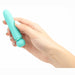 A hand holds a Jaguar 10-Function Rechargeable Silicone Super-Charged Bullet, teal and cylindrical with a rounded tip. A metallic band adds elegance. White-painted nails contrast against the plain background.