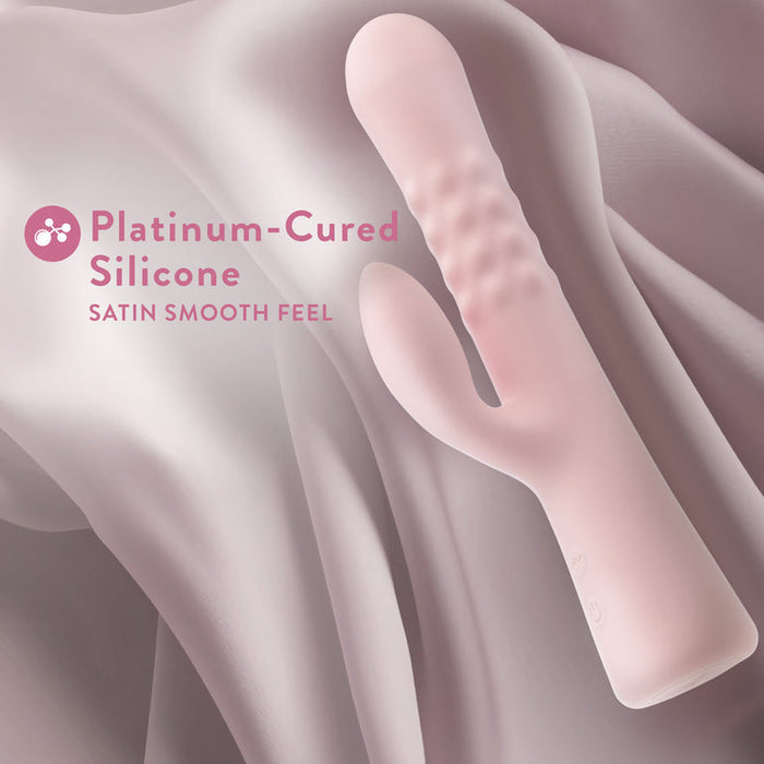 A pink silicone Jaymie G-Spot Rabbit with Pulsating Beads from Blush featuring a ribbed texture and a curved arm is displayed against a soft, wavy backdrop. The text reads "Platinum-Cured Silicone" and "Satin Smooth Feel," highlighting its dual stimulation capabilities.