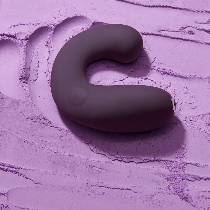 A dark purple, C-shaped electronic device, the Je Joue G-Kii Flexible Clitoral & G-Spot Vibrator by Je Joue, rests on a textured surface covered in lavender-colored powder, creating a contrasting and visually interesting background. This USB rechargeable vibrator features Je Joue's rumbly motor, and the powder appears slightly disturbed where the device is placed.