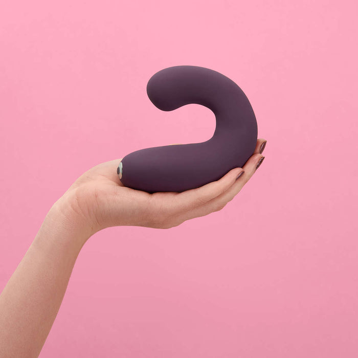 A hand holds the Je Joue G-Kii Flexible Clitoral & G-Spot Vibrator in a rich purple hue, its curved shape featuring a slightly bulbous end, set against a pink background. The sleek design indicates it could be an adjustable vibrator with the renowned Je Joue rumbly motor. The person's nails are painted in a neutral tone.