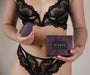 A woman dressed in black lace lingerie holds a Je Joue Mimi Clitoral and External Vibrator - Purple, an egg-shaped item known for its exceptional clitoral stimulation, in her right hand and a purple box labeled "Je Joue" in her left hand. The background is neutral and uncluttered.