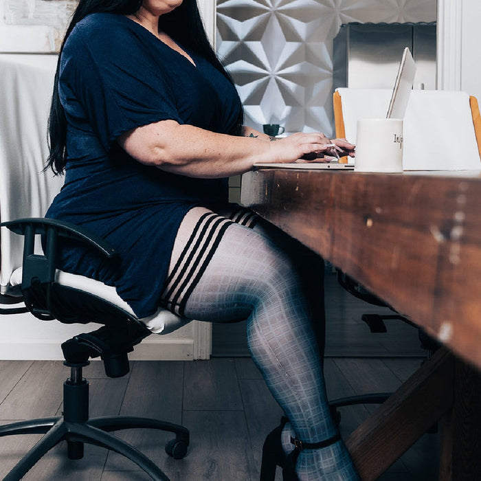 A person with long dark hair and a blue dress is seated at a wooden desk, typing on a laptop. They are wearing Kix'ies Debbie Navy Blue Plaid Thigh Highs (sizes A-D) with black heels. The background features a geometric-patterned wall and a white chair.