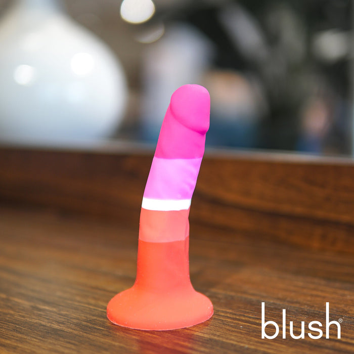 The Lesbian Pride 5.5 Inch Silicone Suction Cup Dildo from Blush, adorned with vibrant pink, red, and purple bands, stands upright on a wooden surface. Made of silky silicone and featuring a suction cup base for hands-free fun, the background is softly blurred with the word "Blush" visible in the bottom right corner.