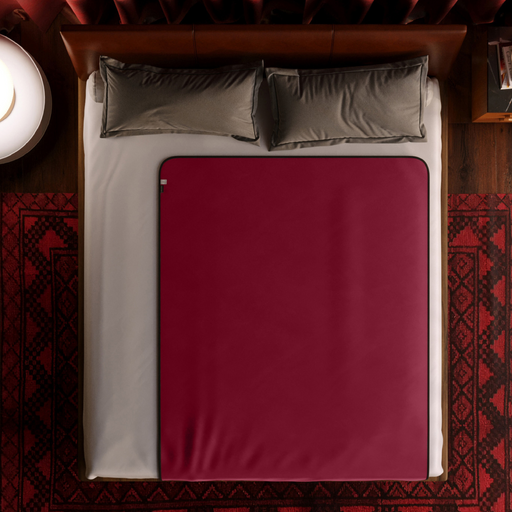 A neatly made bed with a burgundy red Liberator Fascinator Throw Travel Sized Velvety Sex Blanket covering half of it, placed in a cozy room. The bed has two gray pillows and a white bedsheet. The luxurious touch of the Liberator throw adds elegance, while the room features a warm ambiance with a patterned red and black rug and a round bedside table with a lamp.