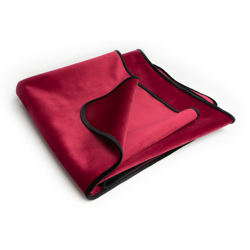 A folded Liberator Fascinator Throw Travel Sized Velvety Sex Blanket in red, featuring a sleek black trim, is neatly placed on a white surface. The blanket's luxuriously soft and velvety texture is prominently visible, along with its rich color, and it also offers moisture barrier protection to ensure you stay comfortable and dry.