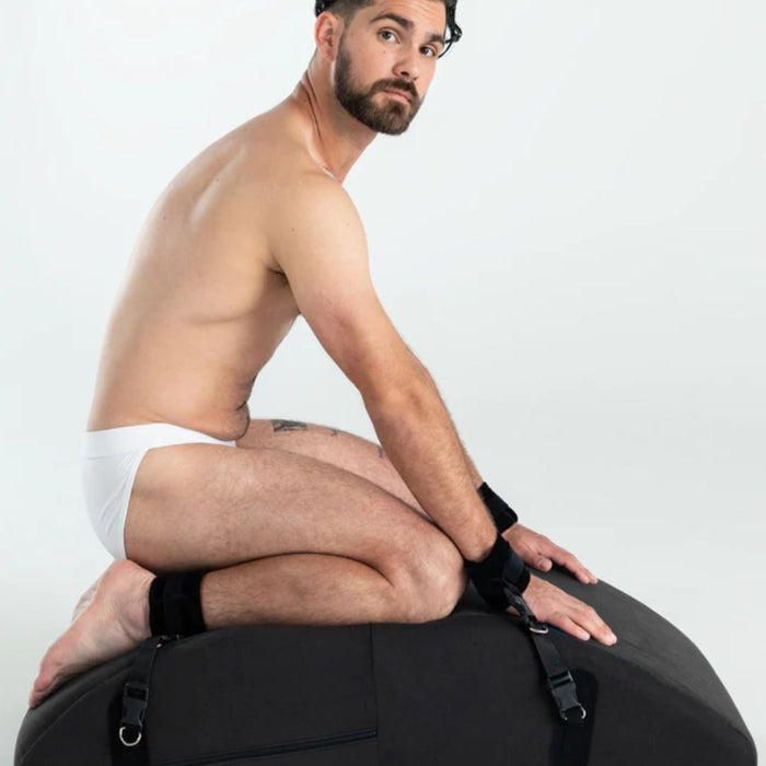 A man with a beard and short hair, wearing only white underwear and black wrist cuffs, is sitting on his knees atop the intimate positioning cushion Liberator Liberator Scoop Rocker Wedge with Cuffs - Black. The background is plain white.