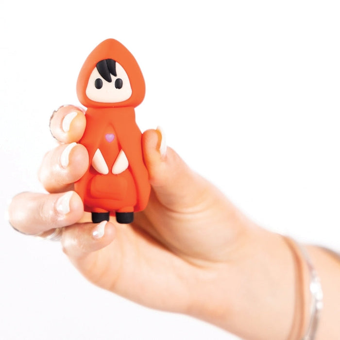 A hand holds a small figurine modeled as the Little Red First Time Discreet Powerful Bullet Vibrator by Natalie’s Toy Box. This red-caped, hooded character has black hair, a white face, and a heart symbol on its chest, complemented by black shoes. Designed to blend seamlessly into any setting, it is set against a plain white background.