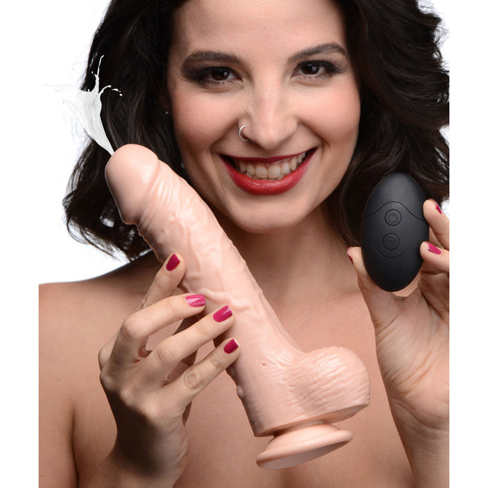 A smiling woman with dark curly hair and a nose ring holds the Loadz 8.5 Inch Vibrating Squirting Dildo with Wireless Remote Control - Vanilla by XR Brands in one hand and a wireless remote control with buttons in the other. The dildo appears to be squirting a white liquid. The woman presents the items closely to the camera, highlighting its 10 modes of vibration.