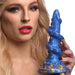 A person with long blond hair and vibrant makeup holds a large, textured, blue and gold Lord Kraken 8.5 Inch Silicone Dildo by XR Brands, resembling a fantasy sculpture. The background is plain white, focusing attention on the person and the intriguing object they are holding.