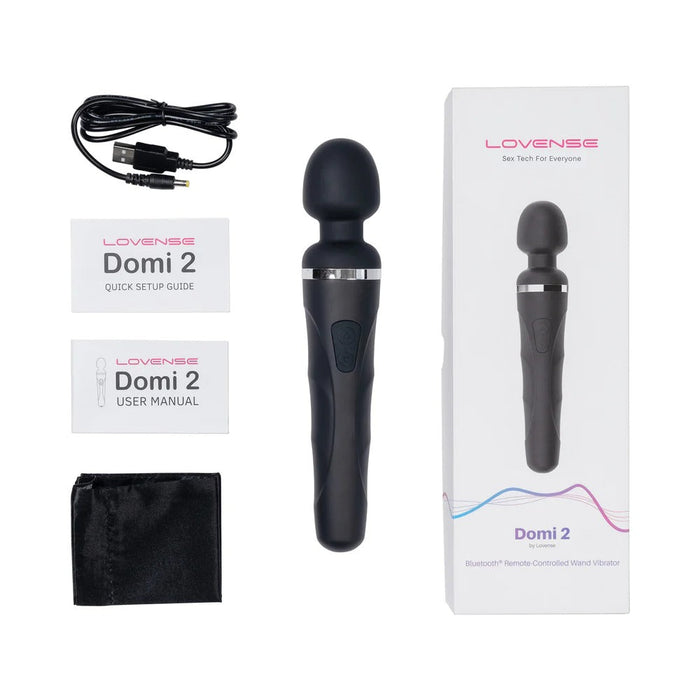 The image showcases the contents of a Lovense Domi 2 Powerful App Controlled Wand Vibrator package, featuring the Bluetooth-compatible wand vibrator, an unopened box, a USB charging cable, a quick setup guide, a user manual, and a storage pouch. The Lovense brand name and product name are clearly visible on the box and documents.