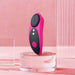 The Lovense Ferri Bluetooth App Controlled Panty Vibrator, featuring a sleek pink and black design with two white LED lights and a power button, is displayed on a clear, circular stand. The backdrop is adorned with pink hues and vertical, translucent lines on the right side. This innovative device from Lovense can be effortlessly controlled via the Lovense app for an enhanced experience.