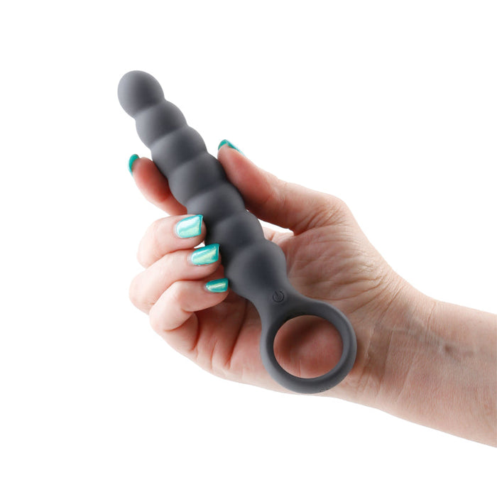 A hand with teal-painted nails holds the NS Novelties Lucent Vibrating Anal Probe with Finger Loop in black. The silicone beaded toy features an ergonomic loop handle with a power button and offers multiple speeds, along with a series of bulbous sections that increase in size towards the handle.