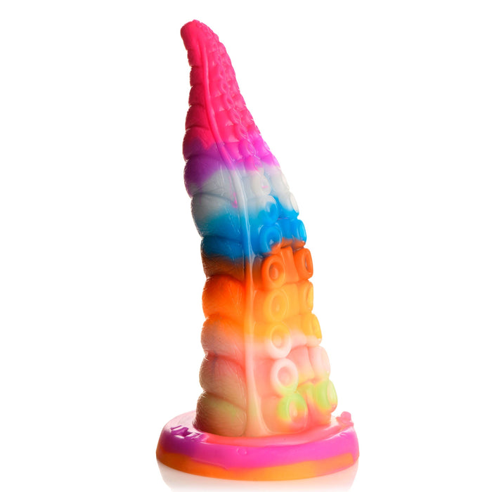 A colorful, silicone tentacle-shaped object standing upright on a white background. The Luminoctopus Glow-in-the-Dark Tentacle Dildo by XR Brands showcases a stunning gradient of vibrant colors, including pink, red, yellow, orange, green, blue, and white. It features textured suction cup-like details and has a flat base that glows in the dark.