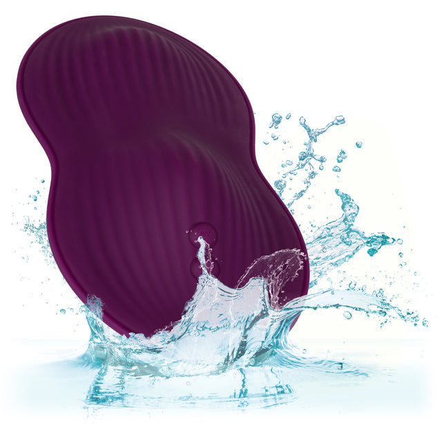 A purple, peanut-shaped device with textured ridges splashes into water, creating droplets and ripples. The plain white background emphasizes the dynamic motion and splash effect of the powerful Lust Dual Rider Remote Control Humping Vibrator by CalExotics.