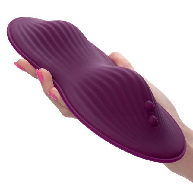 A hand with pink-painted nails is holding a purple, elongated, textured silicone dual motor vibrator from CalExotics, known as the Lust Dual Rider Remote Control Humping Vibrator. The vibrator has two small buttons on one end and is roughly shell-shaped with ripples across its surface, indicating that it's waterproof.