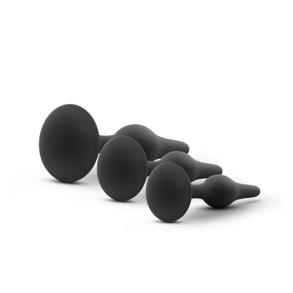 Four black, satin-smooth silicone anal plugs of varying sizes are neatly arranged in a line from largest to smallest. The Luxe Beginner 3 Plug Anal Trainer Set by Blush features plugs with a bulbous end for comfortable insertion and a flared base for added safety. The image has a plain white background.