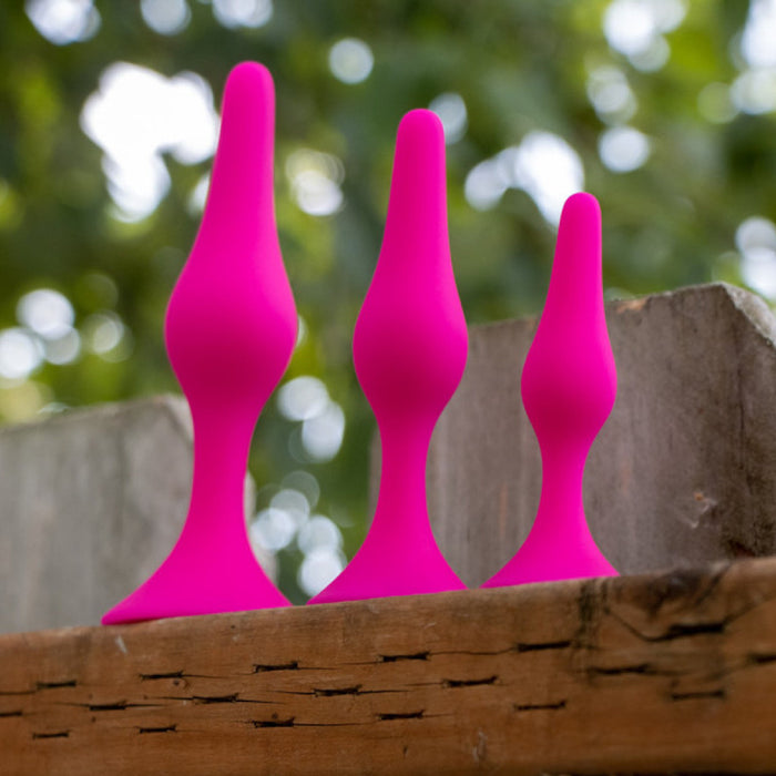 Three pink silicone butt plugs from the Luxe Beginner 3 Plug Anal Trainer Set by Blush are lined up from largest to smallest on a wooden surface. The background features blurred green foliage, suggesting an outdoor setting. Made from body-safe silicone, each plug boasts tapered heads for easy insertion.