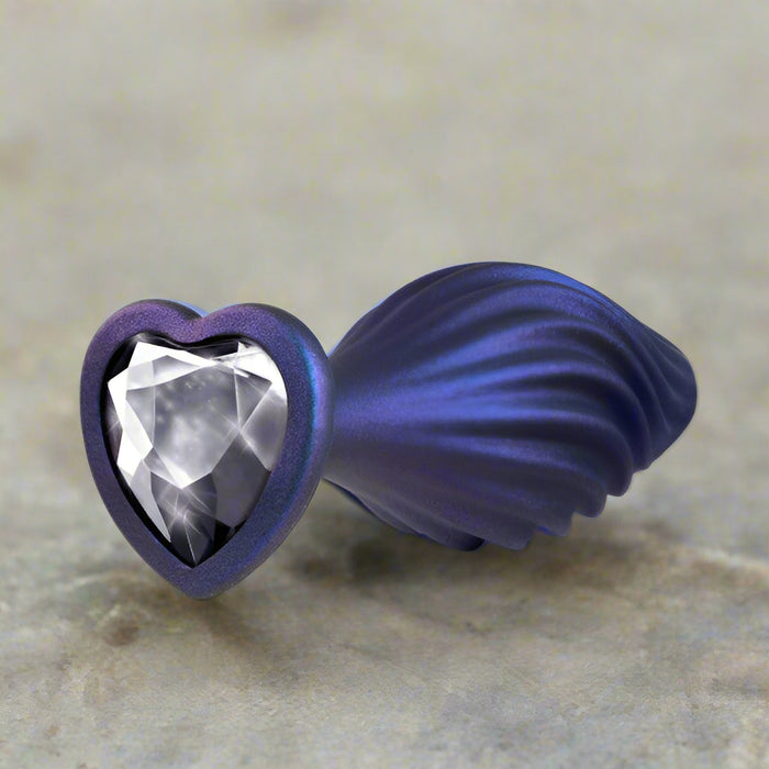 The Matrix Swirl Bling Butt Plug with Sparkly Heart Base by Blush features a dark blue, metallic, heart-shaped base adorned with a large clear faceted gemstone in the center. Its decorative and ornate appearance is enhanced by a seashell-like design on one side, making it both beautiful and functional.