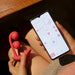 Someone is holding a red Melody Pressure Sensing App-Controlled Wearable Vibrator by Honey Play Box in one hand and a smartphone in the other. The smartphone screen displays an app with various controls and settings for the toy. They are seated on a wooden surface.