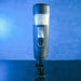 Against a blue background, the Message in a Bottle Thrusting Spinning Stroker with Voices & Mount from Evolved Novelties is showcased on a compact black stand. The sleek, cylindrical design features an illuminated control panel on the front, displaying various buttons and indicators to offer 7 speeds and functions for optimal performance.