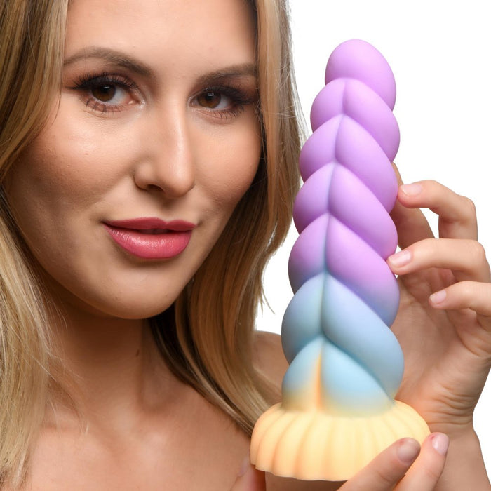 A woman with blonde hair smiles while holding the Mystique Thick 8 Inch Silicone Unicorn Dildo by XR Brands, a whimsical, pastel-toned, spiral-shaped object. The silicone fantasy dildo features a gradient of purple, blue, and orange hues and has a scalloped base. The white background emphasizes the woman and her eye-catching accessory.
