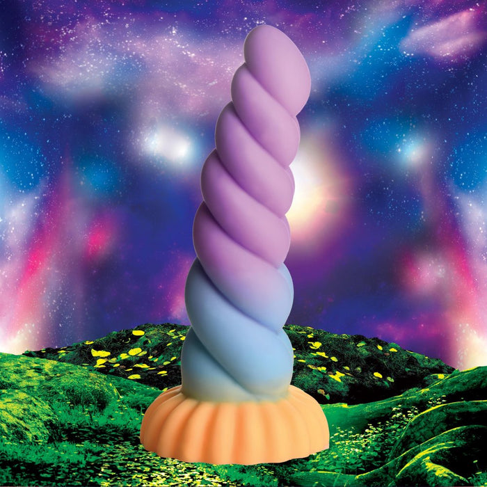 The Mystique Thick 8 Inch Silicone Unicorn Dildo by XR Brands, a spiral, horn-shaped, multicolored sex toy with an orange base, stands on a vibrant green landscape. The background features a bright, surreal sky with glowing, colorful clouds and cosmic patterns, evoking a world of fantasy roleplay.