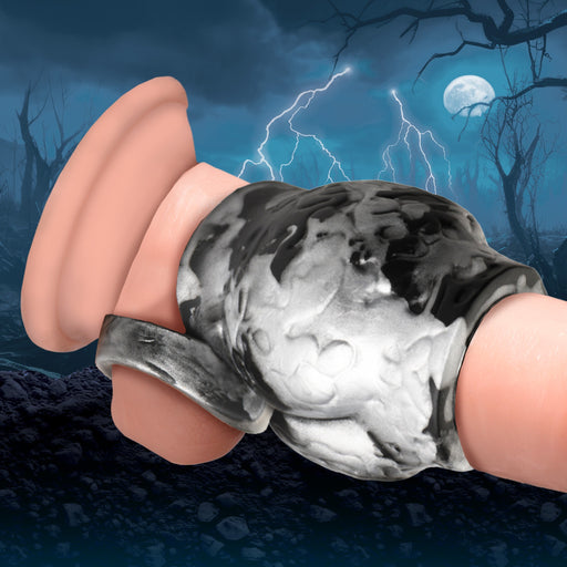 The Night Prowler Alpha Wolf Knot Girth Enhancer with Ball Strap by XR Brands is crafted from body-safe silicone and showcases a striking black and white marbling. In the promotional image, it is set against a dark, ominous background with a full moon, lightning, and barren trees, perfectly conveying a spooky and haunted atmosphere.