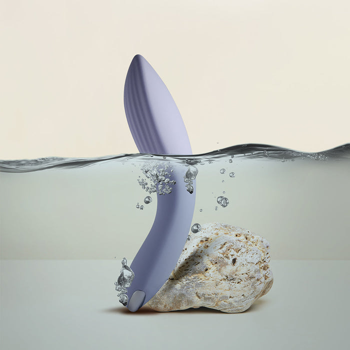 The Niya N2 Powerful Textured Solo and Couples Vibrator by Rocks Off, a sleek purple leaf-shaped device with a powerful motor, is partially submerged in water, its lower part touching a rock. Water droplets adorn the object as bubbles rise to the surface, hinting at movement. The background remains minimalistic and light-colored.