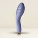 The Niya N2 Powerful Textured Solo and Couples Vibrator by Rocks Off, in a soothing lavender hue, stands upright against a soft, neutral backdrop. Featuring a sleek, curved design with ribbed texture along its surface and equipped with a powerful motor, this vibrator also has a small water drop icon near its slightly reflective base.