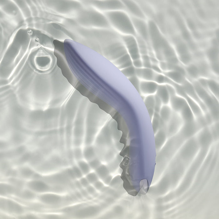 A light purple, curved Niya N2 Powerful Textured Solo and Couples Vibrator from Rocks Off with a ribbed underside is partially submerged in clear water, creating soft ripples and light reflections. Phthalate-free, it rests calmly in the still water, casting gentle shadows on the bottom surface.