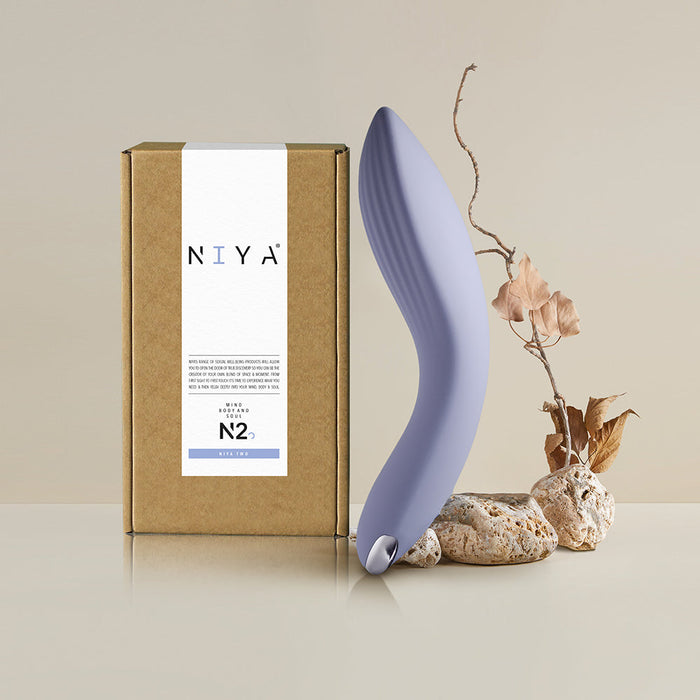 A sleek, blue personal massager with a powerful motor is displayed next to its minimalist, brown packaging box labeled "Rocks Off" and "Niya N2 Powerful Textured Solo and Couples Vibrator." The ergonomically curved massager sits on a reflective surface, accompanied by decorative rocks and dried foliage in the background.