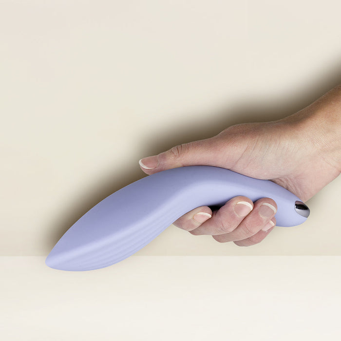 A hand holding the Niya N2 Powerful Textured Solo and Couples Vibrator by Rocks Off—a sleek, pale lavender-colored device with an ergonomic curve and a small metallic button at the base—set against a beige background. The vibrator boasts a powerful motor and exudes modernity.