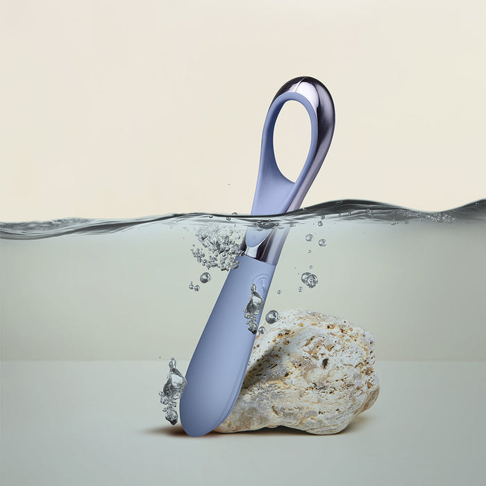 A sleek, modern spoon partially submerged in water, with bubbles rising around it. The spoon's handle is designed with a loop reminiscent of the Niya N3 Easy Grip Finger Loop Bullet Vibrator by Rocks Off, and it is sticking out of a small, light-colored rock positioned at the bottom of the water. The background is plain beige.