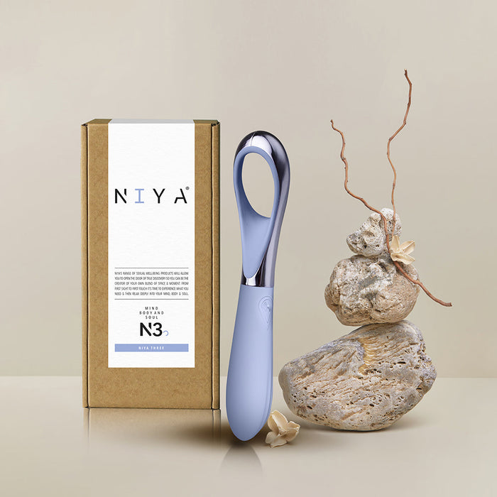 A sleek, blue Niya N3 Easy Grip Finger Loop Bullet Vibrator with a chrome handle is displayed next to its minimalist brown box packaging, which features a label and the brand name "Rocks Off." Set against a neutral background with decorative rocks and a dried plant branch for accent, this waterproof device ensures durability and style.