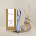 A sleek, blue Niya N3 Easy Grip Finger Loop Bullet Vibrator with a chrome handle is displayed next to its minimalist brown box packaging, which features a label and the brand name "Rocks Off." Set against a neutral background with decorative rocks and a dried plant branch for accent, this waterproof device ensures durability and style.