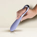 A hand is gently holding the Rocks Off Niya N3 Easy Grip Finger Loop Bullet Vibrator, featuring a purple silicone body with a sleek silver handle, against a cream-colored background. The ergonomic design of the vibrator is tailored for optimal clitoral stimulation, held delicately between the thumb and fingers.