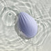 A light purple, shell-shaped device, resembling a modern sleek gadget, floats on the surface of clear, gently rippling water. Crafted from silky silicone, the Niya N4 Palm Held Powerful Textured Egg Vibrator by Rocks Off boasts a smooth, curved design and features a subtle drop-shaped logo on its top surface.