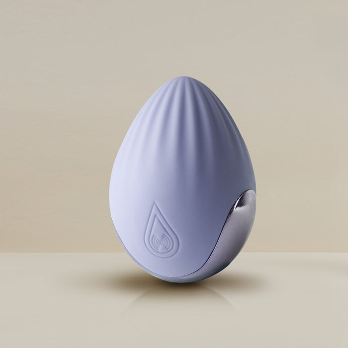 A palm-held device from Rocks Off, the Niya N4 Palm Held Powerful Textured Egg Vibrator, is egg-shaped and light purple with a smooth texture and a ripple pattern on top. It features a teardrop logo with an embedded wireless symbol. Made of silky silicone, this vibrator offers a powerful 10-function motor and rests on a shiny, curved metallic stand against a neutral beige background.