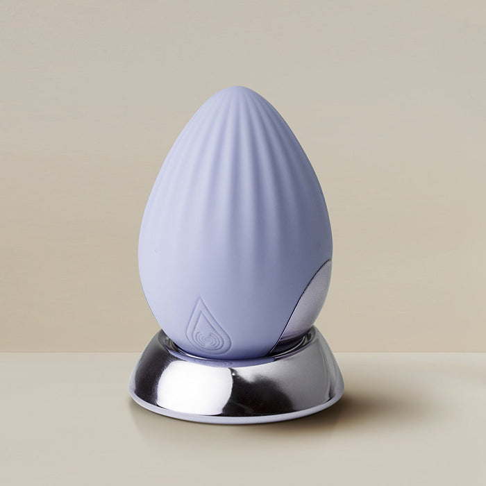 The Niya N4 Palm Held Powerful Textured Egg Vibrator by Rocks Off, a pale lavender silicone device with a soft, ridged texture, rests on a shiny silver base against a neutral background. This palm-held vibrator features a 10-function motor and has a small, embossed water droplet symbol near its base.