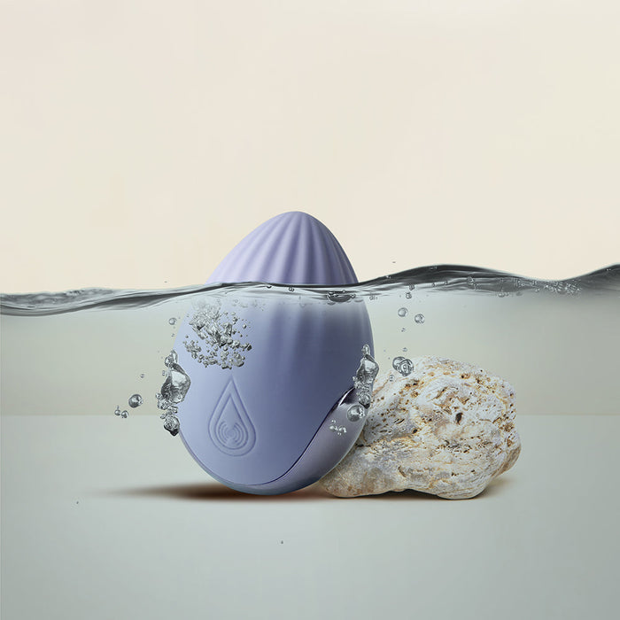 An egg-shaped, light purple waterproof device partially submerged in water, with air bubbles rising to the surface. It sits on a small piece of coral. The silicone Niya N4 Palm Held Powerful Textured Egg Vibrator by Rocks Off features a 10 function motor. The background is neutral, emphasizing the aquatic theme.