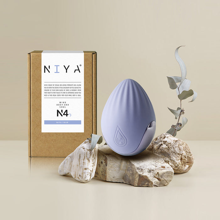 A stylish, egg-shaped palm-held vibrator from Rocks Off, featuring a smooth lavender silicone surface and adorned with a droplet-like logo, is showcased on an arrangement of stones next to a leafy branch. Behind it sits a cardboard box labeled "NIYA" with product details and the model number "N4.