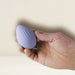 A hand gently cradles the Niya N4 Palm Held Powerful Textured Egg Vibrator from Rocks Off, a small, lavender-colored device with a smooth, textured surface and a faint logo near the bottom. This vibrator boasts a 10-function motor and is conveniently USB rechargeable. The background is a plain, light beige color.